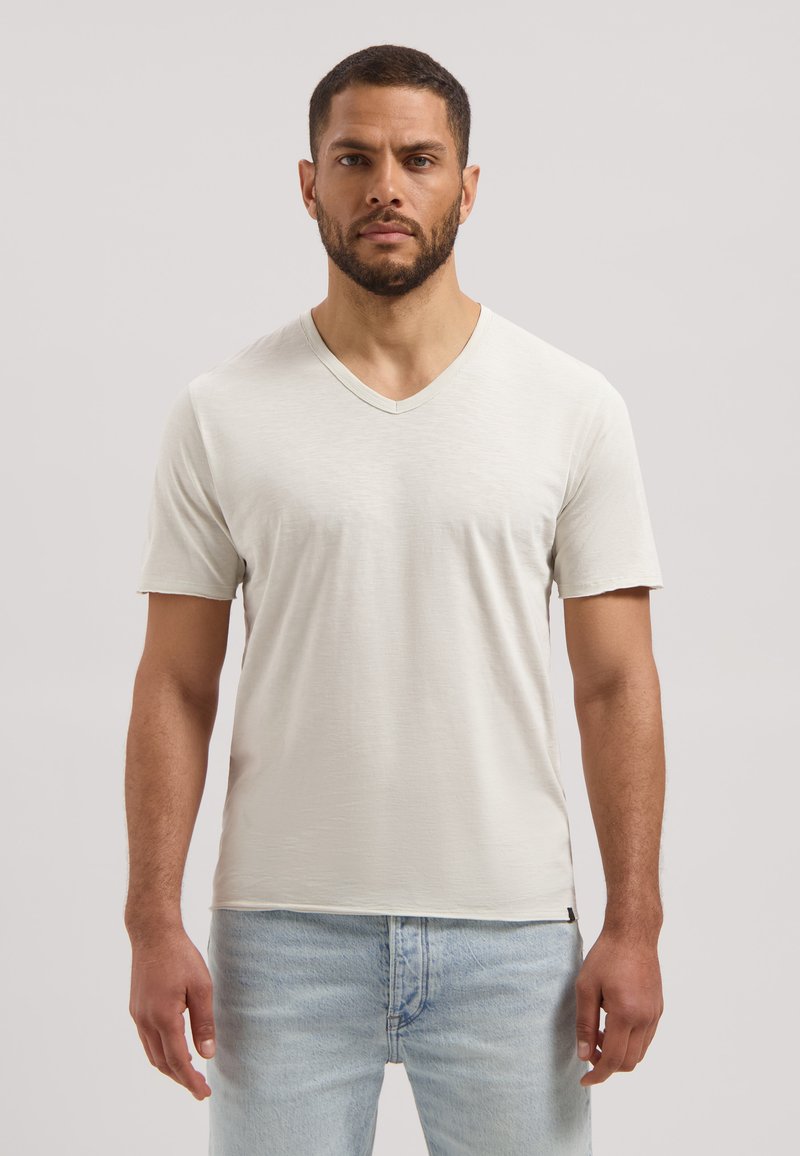 Homme aux cheveux courts et foncés avec une barbe, portant un T-shirt blanc uni à col en V et un jean bleu clair, debout devant un fond uni.