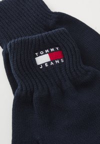 Námornícke pletené rukavice s rebrovaným nátepníkom, na ktorom sa nachádza obdĺžnikové logo Tommy Jeans vyšitá v bielej, červenej a modrej farbe.