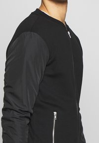 Chaqueta bomber negra que presenta un cuerpo de punto con mangas de nailon negras lisas, una cremallera frontal y bolsillos laterales con detalles de cremallera brillantes.