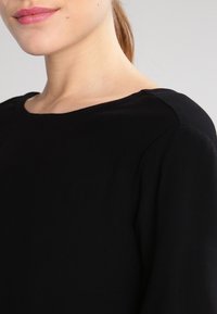 Svart långärmad topp med vid halsringning, slät textur och subtil sömdekor längs axlarna. Enkel och mångsidig design.