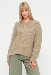 Mladá žena s rovnými blond vlasmi, oblečená v béžovom hrubom pletenom cardigan s gombíkmi a vo voľných zelených nohaviciach, na jednoliatom pozadí.