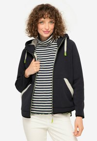 Felpa con zip color navy con fodera grigia, abbinata a un dolcevita a righe grigie e navy. Presenta due tasche laterali e dettagli della zip in verde.