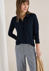 Femme portant une chemise à manches longues bleu marine et un pantalon gris, tenant une veste beige dans sa main droite, debout devant un fond neutre.
