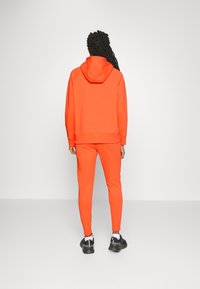 Orange hoodie och matchande joggingsbyxor i ett mjukt material. Har en huva och ribbade mudd, tillsammans med svarta sportskor.