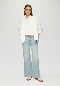 Weißes langärmeliges Button-up-Hemd über einem weißen T-Shirt, kombiniert mit hellblauen weitgeschnittenen Jeans und beigen sowie cremefarbenen Sneakers.