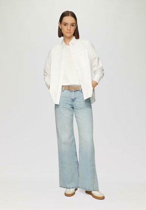 Weißes langärmeliges Button-up-Hemd über einem weißen T-Shirt, kombiniert mit hellblauen weitgeschnittenen Jeans und beigen sowie cremefarbenen Sneakers.