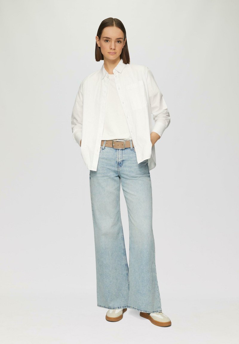 Weißes langärmeliges Button-up-Hemd über einem weißen T-Shirt, kombiniert mit hellblauen weitgeschnittenen Jeans und beigen sowie cremefarbenen Sneakers.