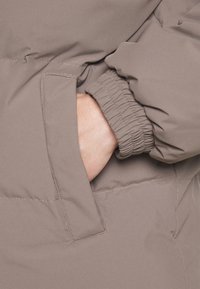 Puffer jacket in light brown fabric, featuring gathered cuffs and a side pocket with visible stitching for secure storage.
