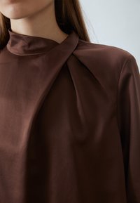 Blusa marrone con un collo alto, caratterizzata da un design intrecciato al colletto. Tessuto liscio, si adagia delicatamente sulle spalle.