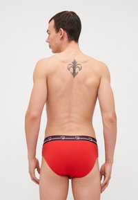 Slip rossi da uomo con un design aderente e una cintura nera a contrasto con strisce bianche e rosse. La vista posteriore mostra un tatuaggio floreale.