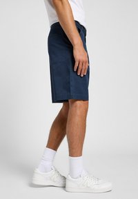 Marineblå shorts lavet af et glat stof, med en standard pasform, sidelommer og en ren kant. Båret med hvide sneakers og sokker.