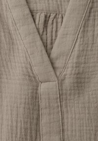 Beige strukturierter Stoff mit genähten Nähten, die eine tiefe V-Ausschnittöffnung bilden, und gerafften Stichen am Halsausschnitt.