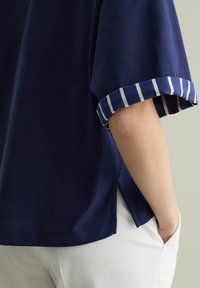 Camiseta azul marino con mangas enrolladas y rayas blancas. Tejido suave, corte relajado, aberturas laterales, combinada con pantalones blancos.
