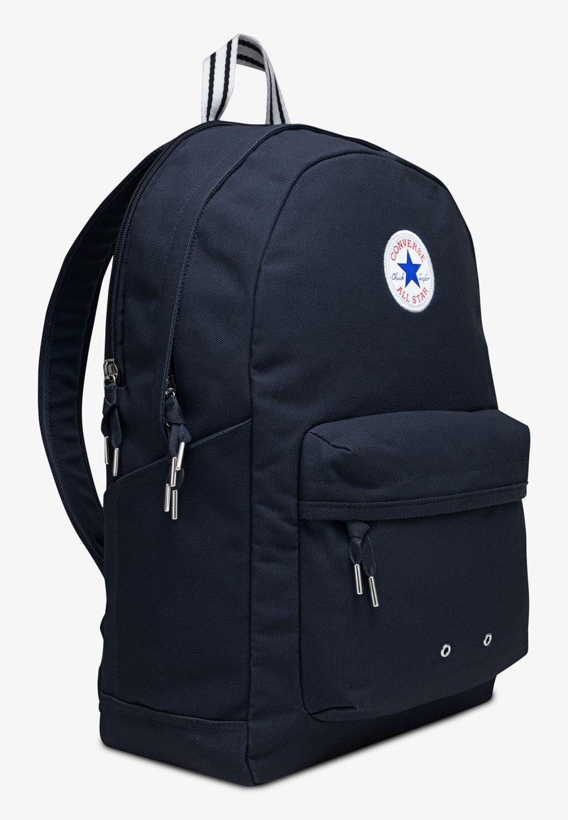 Mochila de tela azul marino con forma redondeada, que cuenta con un bolsillo frontal, doble cremallera y un parche de logo circular. Incluye un asa superior a rayas.