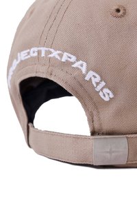 Gorra de béisbol beige con "PROJECTXPARIS" bordado en blanco. Presenta una tela texturizada, correa ajustable y herrajes de metal.