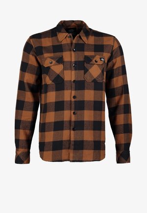 Homme portant une chemise à carreaux marron et noire sur un t-shirt blanc, un jean skinny bleu clair, des baskets montantes rouges, et portant un sac à bandoulière noir.