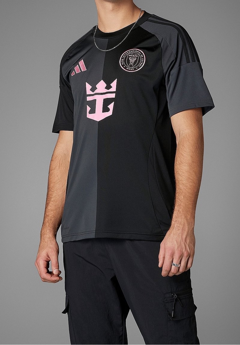 Homme portant un maillot de football noir et gris foncé avec des logos roses, incluant un emblème de couronne et un badge circulaire pour Club Internacional Futbol Miami.