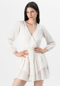 Modella con lunghi capelli scuri posa con confidenza, indossando un vestito color crema con scollatura a V profonda e gonna a strati, su uno sfondo neutro.