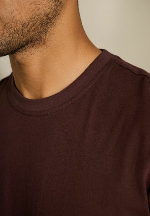 Prim-plan al gâtului și bărbiei unui bărbat cu stubble scurt, purtând un tricou cu guler rotund de culoare maro închis, pe un fundal deschis, uniform.