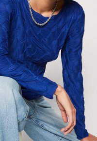 Blå långärmad topp i texturerad tyg med vågmönster. Har rund halsringning och avslappnad passform. Matchas med ljusblå jeans.