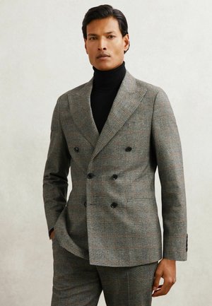 Dwurzędowy szary garnitur w kratę z teksturowanej tkaniny, ozdobiony pomarańczowymi liniami i czarnymi guzikami, noszony na czarnym golfie.