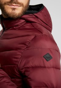 Burgundská péřovka s prošívanou texturou, černým podšívkou kapuce a černou nášivkou loga. Detailní záběr na oblast ramene ukazuje detaily šití.