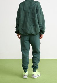 Osoba nosící tmavě zelenou prošívanou mikinu s vystouplými logy swoosh, odpovídající tepláky a neonově zelené tenisky, stojící na zelené podlaze.