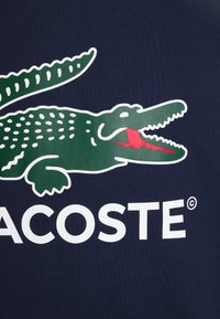 T-shirt in cotone blu navy con logo di coccodrillo verde con lingua rossa e testo bianco "Lacoste" sottostante. Texture liscia, design casual.