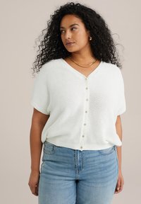 Cardigan tricoté blanc à manches courtes avec un col en V et une fermeture à boutons ; présente un motif texturé et un ourlet côtelé. Porté avec un jean bleu clair.