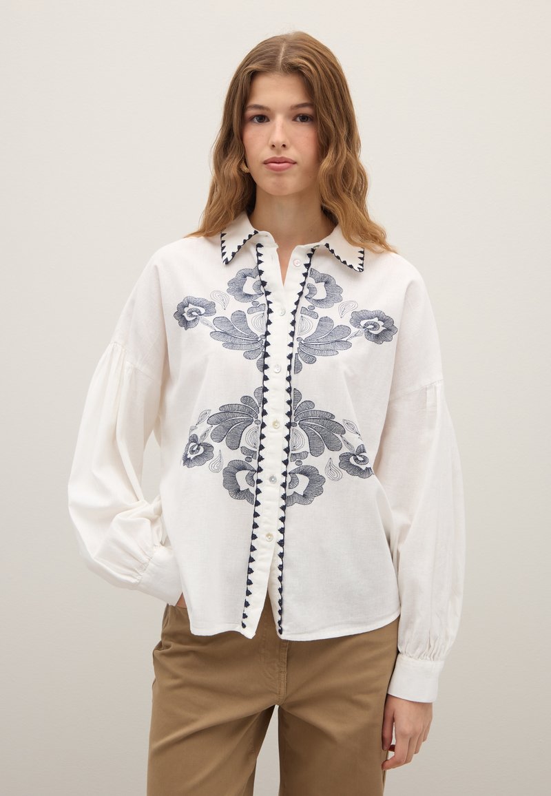 White embroidered blouse with blue floral patterns, button-down front, and scalloped collar. Features long sleeves with gathered cuffs.