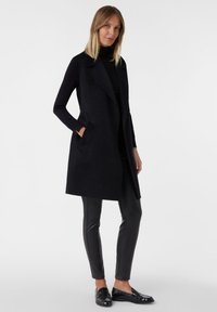 Schwarzer langärmeliger Cardigan aus strukturiertem Stoff mit einem Schal-Kragen und seitlichen Taschen, kombiniert mit glänzenden schwarzen eng anliegenden Hosen und Slippern.