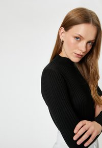 Jersey de punto negro con cuello alto y mangas largas, que muestra un patrón texturizado. La modelo tiene el cabello largo y castaño y está posando con los brazos cruzados.