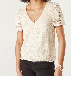 Mujer con blusa de encaje crema de manga corta y escote en V, pantalones negros, collares en capas y pulseras en la muñeca derecha.