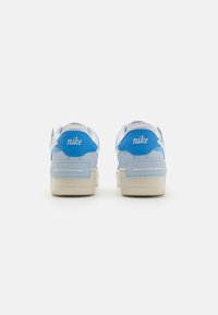 Tênis Nike com um cabedal em couro branco, detalhes em azul claro, colarinho acolchoado e sola em borracha texturizada. A marca está bordada em branco.