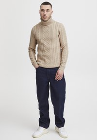 Beige kabelstrikket turtleneck-genser med ribbede mansjetter, sammen med mørkeblå jeans og hvite sneakers med gule detaljer.
