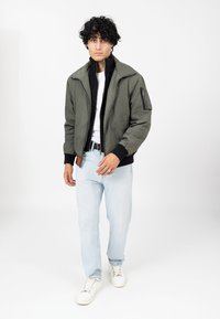 Veste bomber vert olive avec poche zippée, poignets et col noirs ; associée à un jean bleu clair et des baskets blanches.
