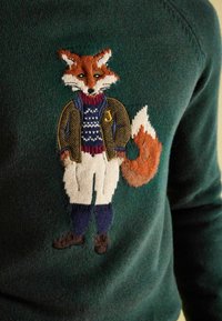 Gestrickter Fuchs in einer braunen Jacke, einem blau gemusterten Pullover, weißen Hosen und braunen Schuhen auf einem dunkelgrünen Pullover.