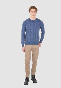 Maglione azzurro a trama a treccia con polsini e vita a coste, abbinato a pantaloni beige e stivali neri, modello in piedi su uno sfondo neutro.