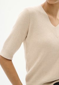 Beige ribbad stickad tröja med V-ringning och korta ärmar, med en mjuk textur och en figurnära silhuett.