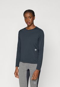 Dlhý rukáv tmavomodré tričko vyrobené z ľahkého materiálu, s okrúhlym výstrihom a malým logom na prednej strane, skombinované so šedými nohavicami.