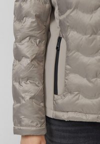 Lichte beige pufferjack met een glanzende textuur, met een ritszak aan de zijkant en een snugger polsmanchet.