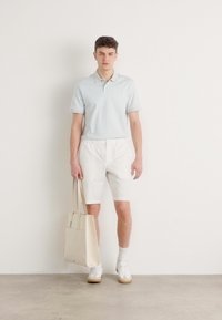 Polo shirt bleu clair, shorts blancs, chaussettes blanches et baskets. Tient un sac fourre-tout beige clair. Matériaux lisses et design simple.