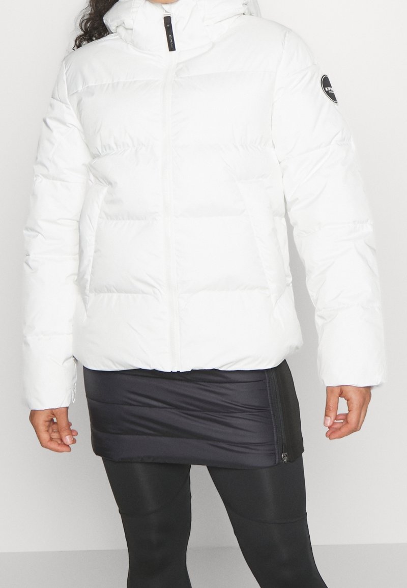 Vit vadderad jacka med dragkedja framtill, hög krage och elastiska muddar. Kännetecknas av sin kviltade design och logotypdetalj på ärmen.