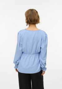 Lyseblå bluse med lange pufærmer, elastisk taljelinje og bølget kant ved halsen. Tekstureret stof med en blød finish.