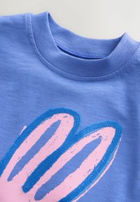 Povečani posnetek modre majice z okroglim izrezom in teksturirano roza-modro grafiko zajčjih ušes na sprednji strani.
