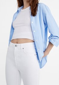 Světle modrá košile s dlouhými rukávy vyhrnutými, nošená přes bílý ribovaný crop top, v kombinaci s bílými džínsy s vysokým pasem.
