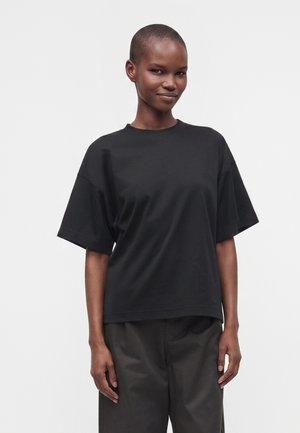 Kvinde med kort hår iført en ensfarvet sort oversized t-shirt og mørke bukser, stående mod en hvid baggrund.