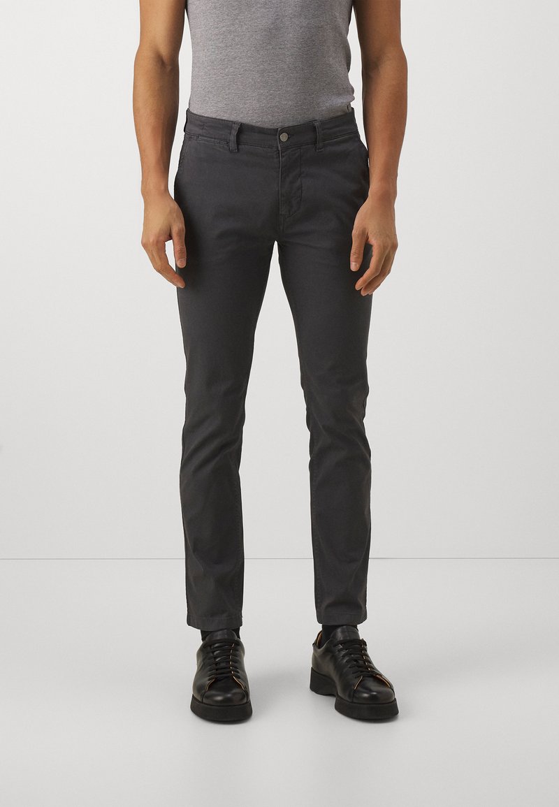 Pantalon en coton gris à coupe slim, avec fermeture à boutons, poches latérales et poches arrière, associé à des chaussures noires à lacets.