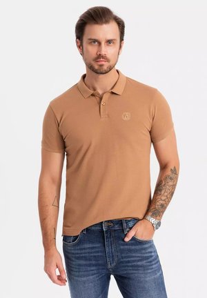 Polo de color beige con collar clásico, mangas cortas y detalle de logo en el pecho, combinado con jeans de mezclilla azules y un reloj.
