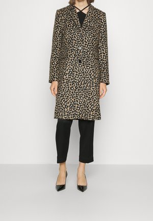 Kobieta ubrana w płaszcz w panterkę do kolan, czarny top z krzyżującymi się ramiączkami, czarne spodnie do połowy łydki oraz czarne szpilki w szpic, stojąca na białym tle.